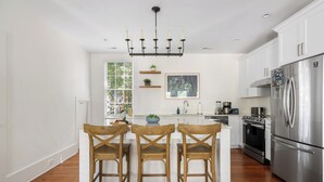 A modern kitchen with white cabinets, a stainless steel refrigerator, wooden chairs, and a chandelier.