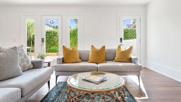 A modern living room with a grey sofa, yellow cushions, a glass coffee table, and a view of greenery through large windows. - Private Pool! | Rooftop Patio | The Athena (Charleston)