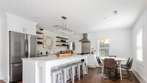 A modern kitchen with a stainless steel refrigerator, white cabinetry, a central island with bar stools, a dining table with chairs, and a large window with blinds. - Private Pool! | Rooftop Patio | The Athena (Charleston)