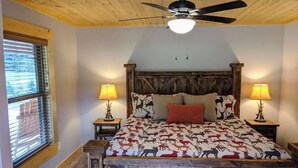 Un dormitorio con techo de madera, una cama con colcha estampada, dos lámparas de noche y un ventanal con persianas.