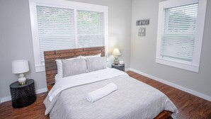 Un dormitorio con cabecera de madera, cama con edredón blanco, dos mesitas de noche con lámparas y una ventana con persianas.