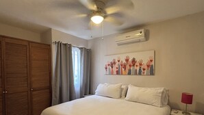 Un dormitorio con cama, ventilador de techo, aire acondicionado, mesita de noche con lámpara y un cuadro de manos en la pared.