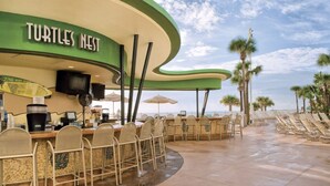 Eine Strandbar mit einem gebogenen, grünen Sonnensegel, Sitzgelegenheiten im Freien und Palmen im Hintergrund.