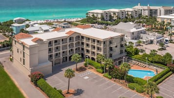 A multi-story hotel with a pool, surrounded by a parking lot and palm trees, overlooking a beach.