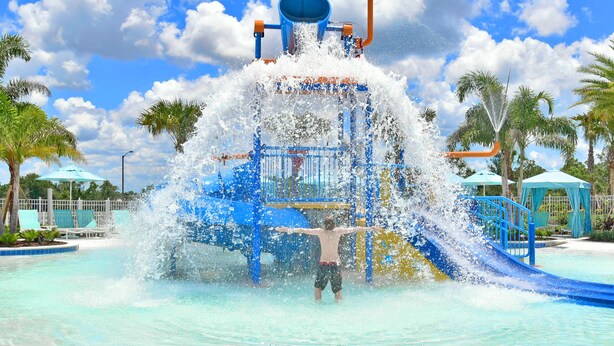 Area giochi acquatici con un bambino in acqua, circondata da palme e cielo sereno.