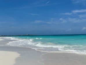 Ein Strand mit klarem türkisblauem Wasser und einer fernen Küstenlinie mit Gebäuden.