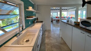 A modern kitchen with a large window, white cabinets, and a view of the outdoors. - Fair Winds: Tropical Beach House, Stunning Ocean View, Pool, Steps to the Beach! (Farmer's Hill)