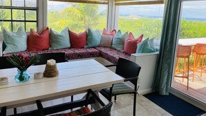 A cozy seating area with colorful cushions, a table with a vase of flowers, and a view of lush greenery and the sea. - Fair Winds: Tropical Beach House, Stunning Ocean View, Pool, Steps to the Beach! (Farmer's Hill)
