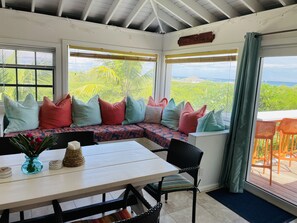 A cozy seating area with colorful cushions, a table with a vase of flowers, and a view of lush greenery and the sea. - Fair Winds: Tropical Beach House, Stunning Ocean View, Pool, Steps to the Beach! (Farmer's Hill)
