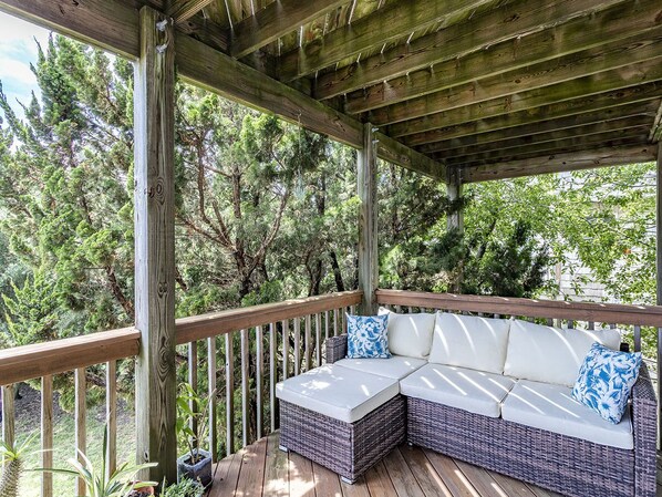 Een overdekt terras met een houten dek, een rieten bank met kussens, en uitzicht op weelderige bomen.