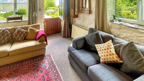 Un salon chaleureux avec un canapé beige, un canapé gris et un tapis à motifs. On y trouve une fenêtre avec des rideaux, un miroir au mur et une porte en bois donnant sur l’extérieur.