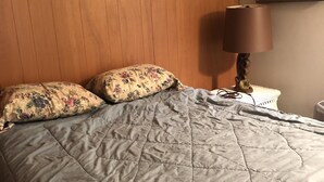 Une chambre avec une tête de lit en bois, une couvre-lit matelassée, des coussins à motifs floraux, une lampe de chevet et un tableau au mur.