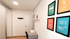 Um quarto pequeno e bem organizado, com escrivaninha, cadeira e cama. Quatro cartazes motivacionais emoldurados estão na parede.