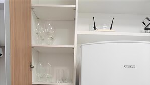 Uma cozinha moderna com geladeira branca, prateleiras abertas com copos e canecas brancas, e uma porta de madeira.