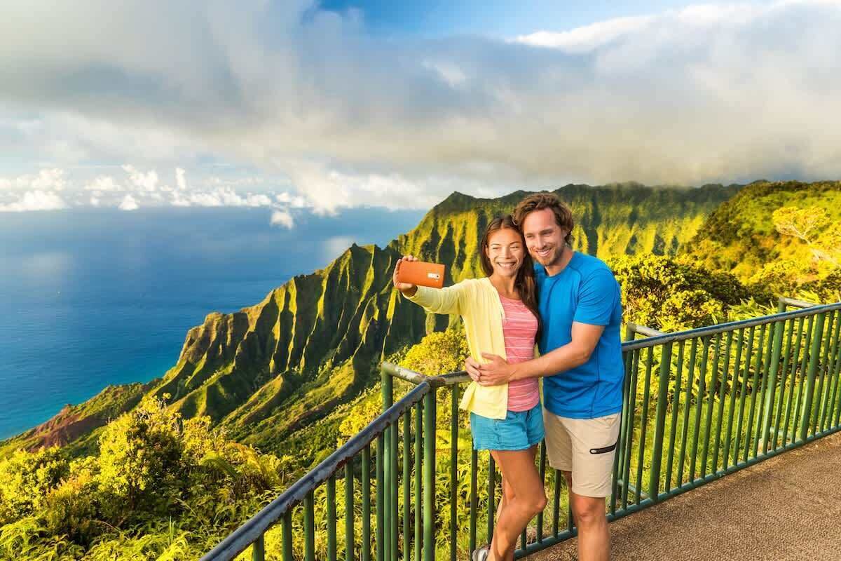 A couple taking a selfie on a scenic overlook with a view of a mountainous coastline.
