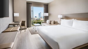 A modern hotel room with a large bed, a desk, a chair, and a sofa. There is a TV mounted on the wall and a window with a view of the city. - AC Hotel by Marriott Raleigh Downtown (Raleigh)