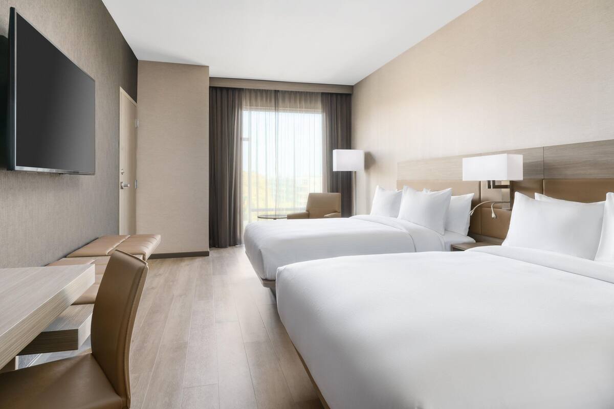 A hotel room with two beds, a desk, a chair, a TV, and a window with curtains.