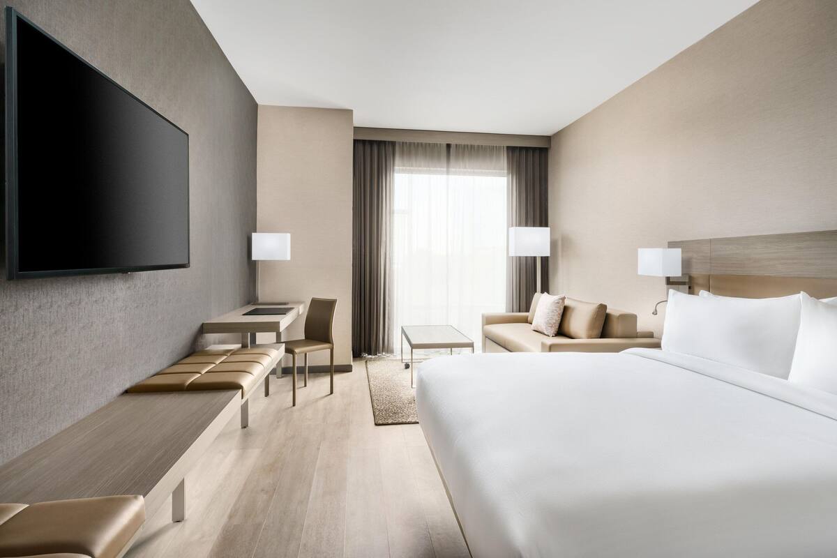 A modern hotel room with a large bed, a desk with a chair, a sofa, and a flat-screen TV mounted on the wall.