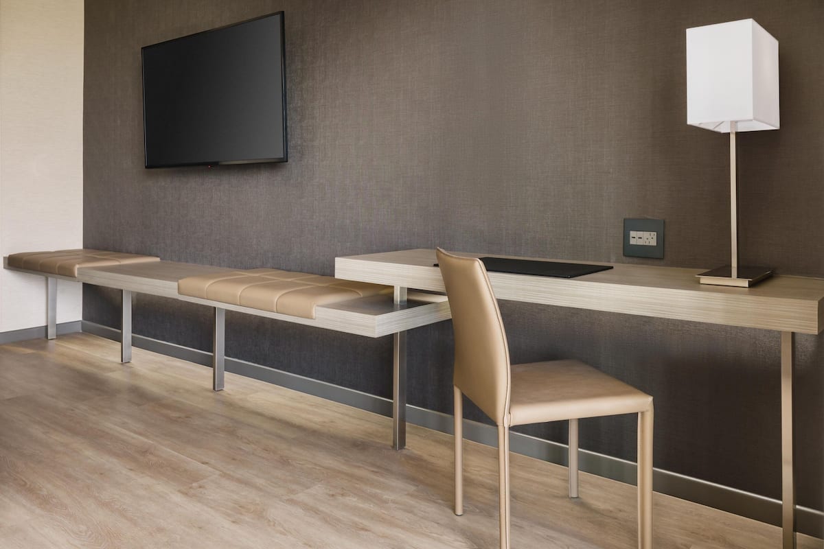 A modern hotel room with a long bench, a desk, a chair, a flat-screen TV, and a floor lamp.