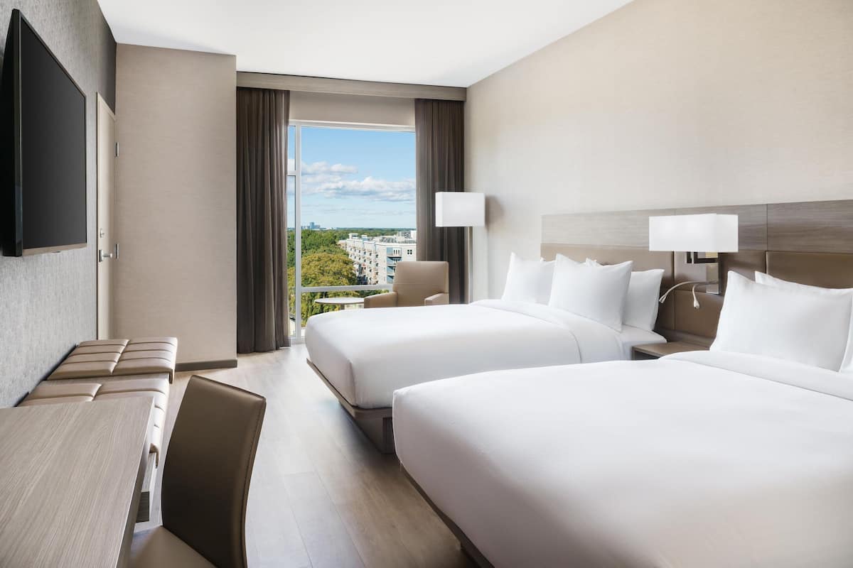 A hotel room with two beds, a desk, a chair, a TV, and a window with a city view.