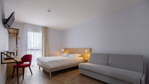 A modern hotel room with a bed, a desk, a red chair, a grey sofa, and a TV mounted on the wall. - The Originals City, Hôtel Le Relais des Deux Mers, Marmande Sud (Samazan)