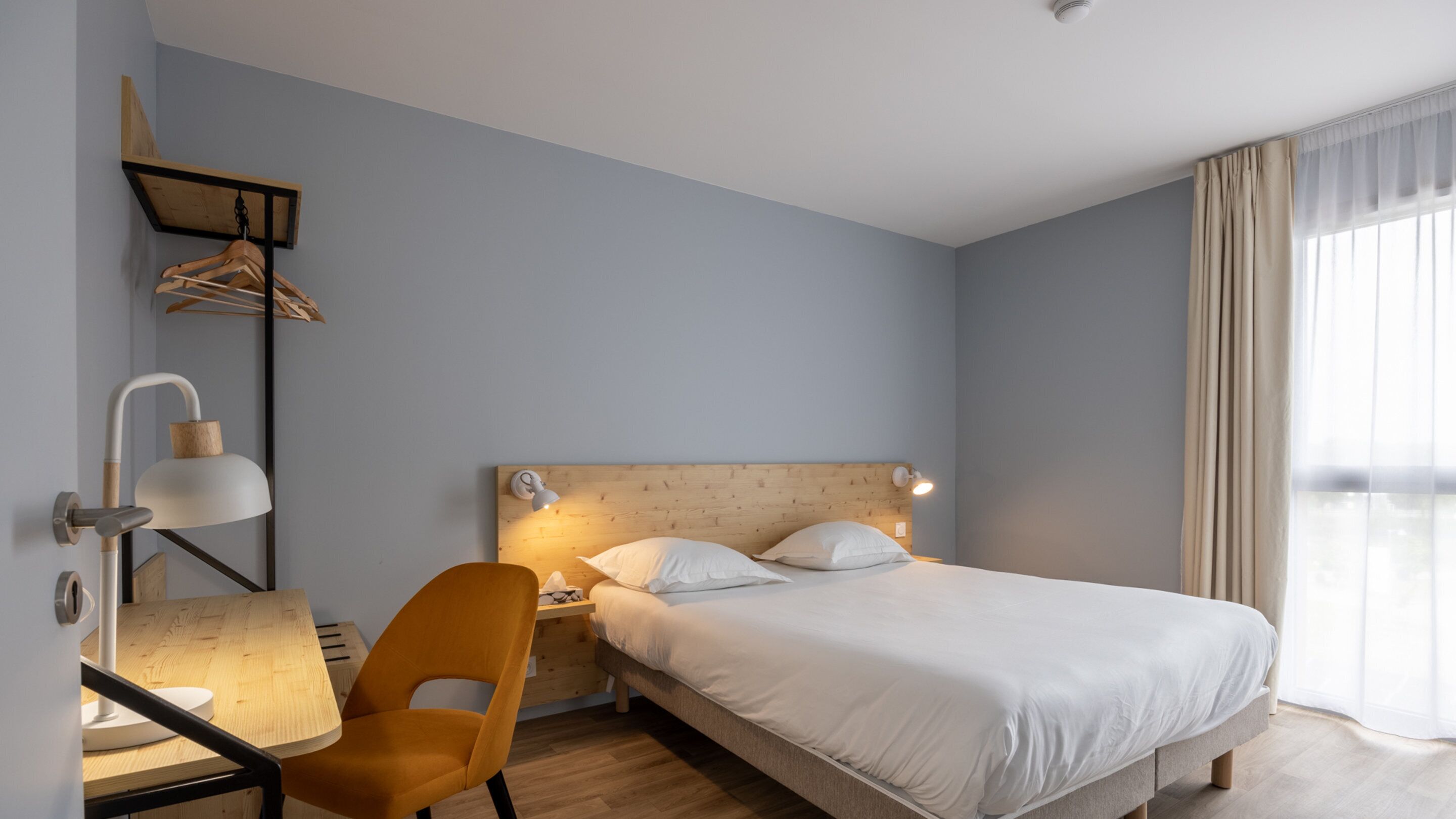 Un dormitorio con cama, escritorio con silla, lámpara de pared y cabecero de madera.