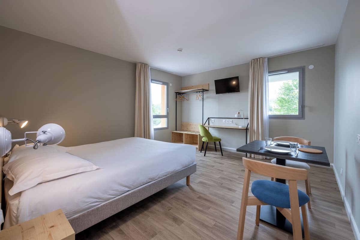 A hotel room with a bed, desk, chair, TV, and windows.