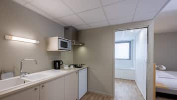 Cocina compacta con microondas, cafetera y lavabo. Una pequeña ventana proporciona luz natural y una puerta da a un dormitorio con cama.