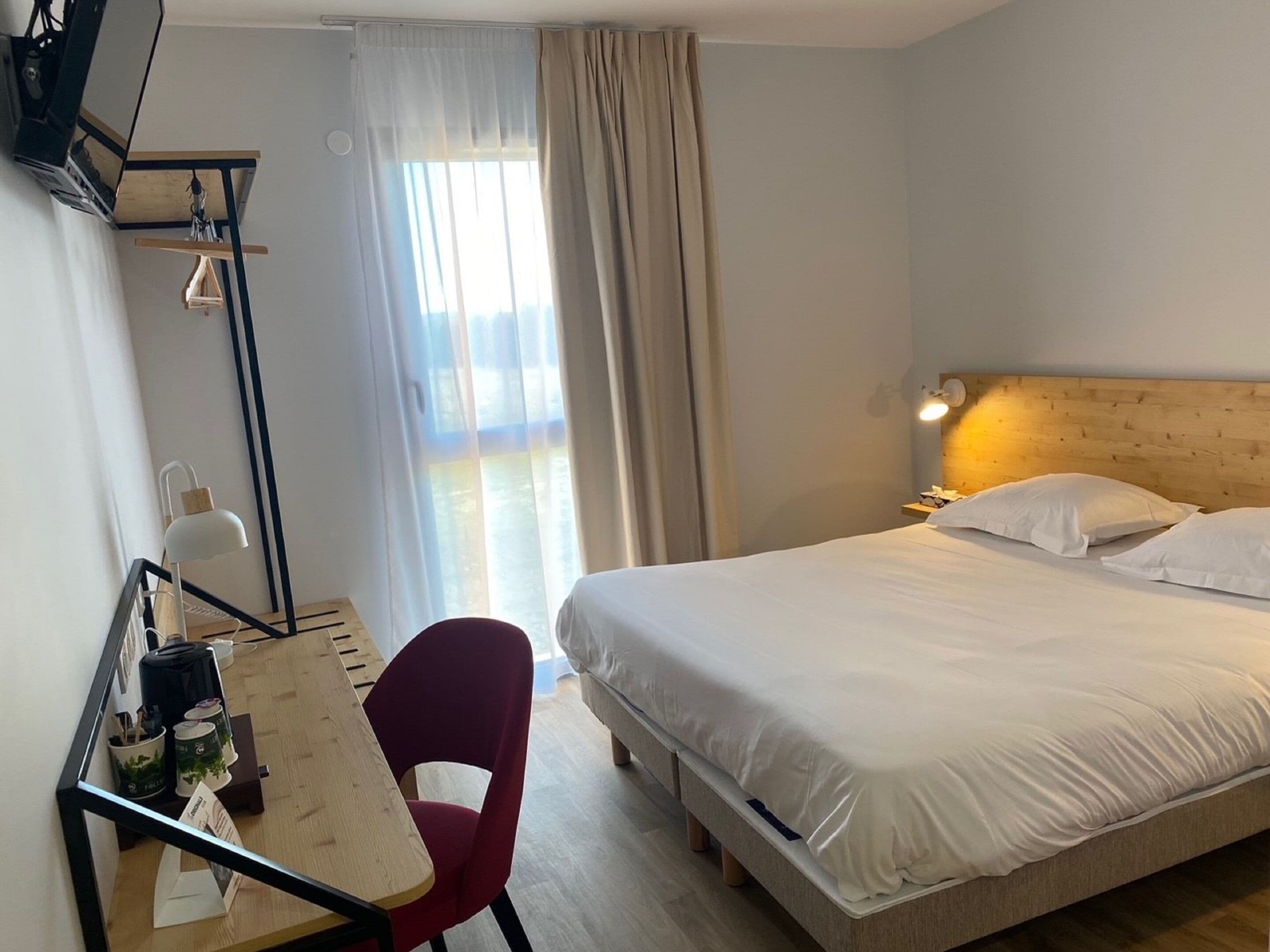 Ett hotellrum med en säng, ett skrivbord med en stol, en lampa och ett fönster med gardiner.