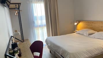 Ett hotellrum med en säng, ett skrivbord med en stol, en lampa och ett fönster med gardiner.