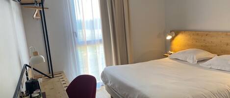 A hotel room with a bed, a desk with a chair, a lamp, and a window with curtains.