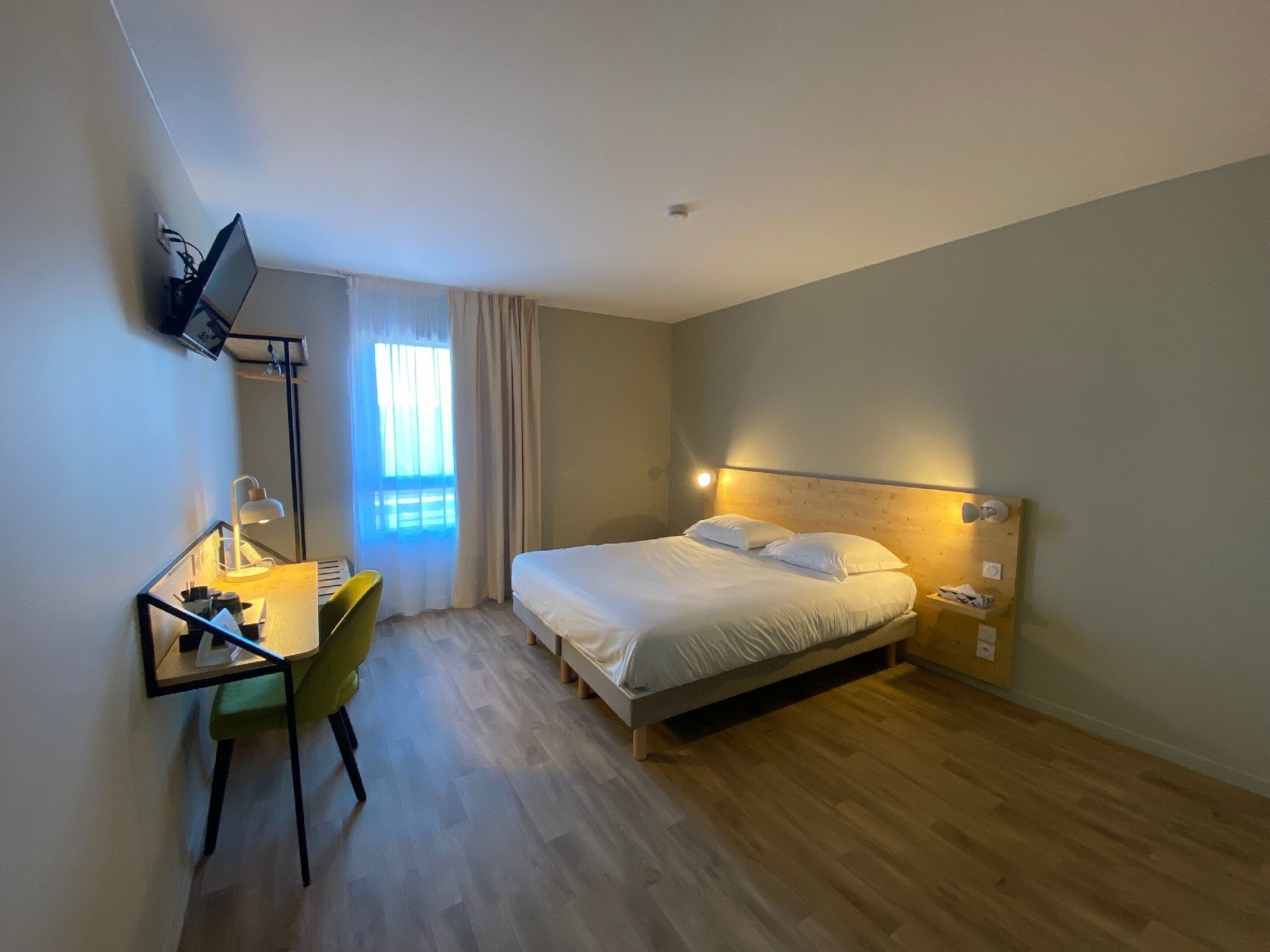 A hotel room with a bed, a desk with a chair, a TV mounted on the wall, and a window with curtains.