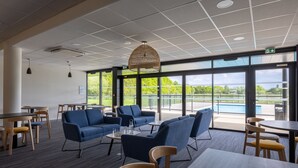 A modern lounge area with blue sofas, wooden tables, and a large window offering a view of a swimming pool and greenery. - The Originals City, Hôtel Le Relais des Deux Mers, Marmande Sud (Samazan)