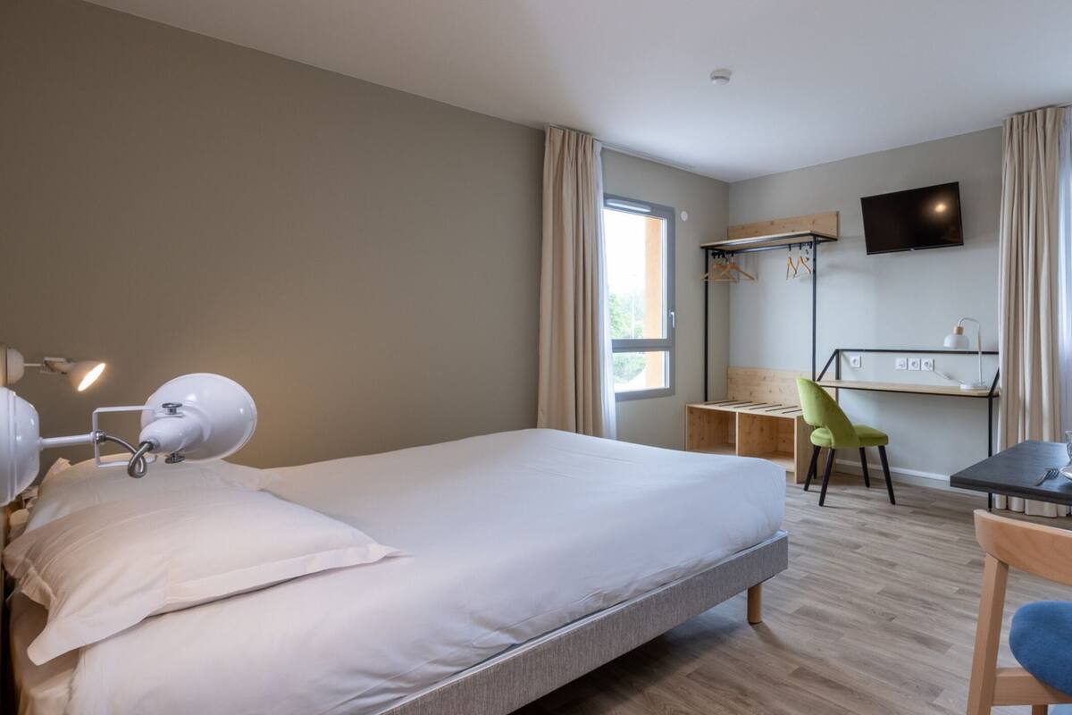A modern hotel room with a large bed, a desk, a chair, and a TV mounted on the wall.