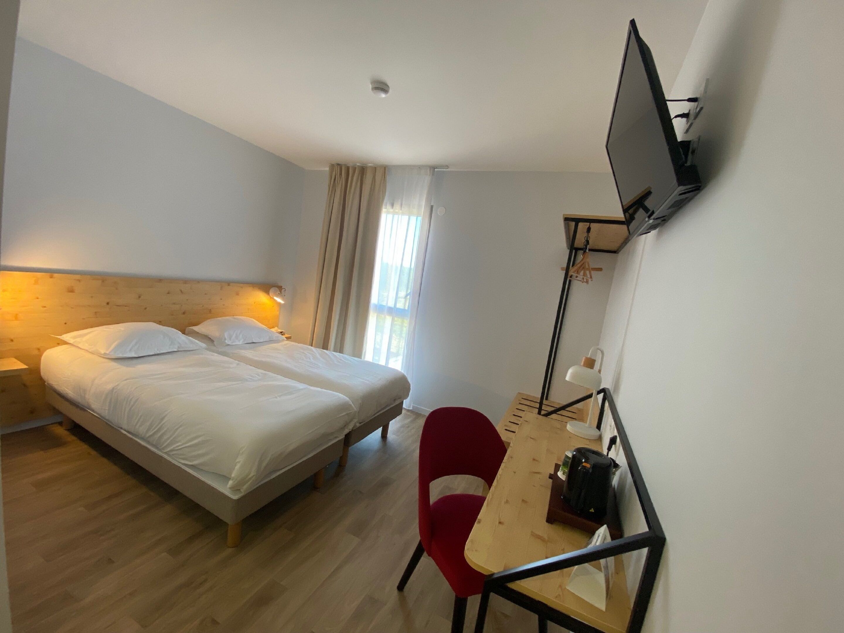 A hotel room with two beds, a desk with a chair, a TV mounted on the wall, and a window with curtains.