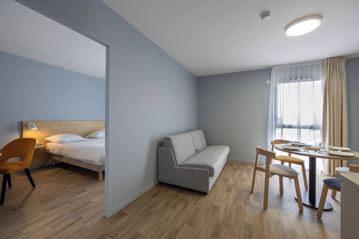 A modern, well-lit room with a bed, sofa, dining table, and chairs.