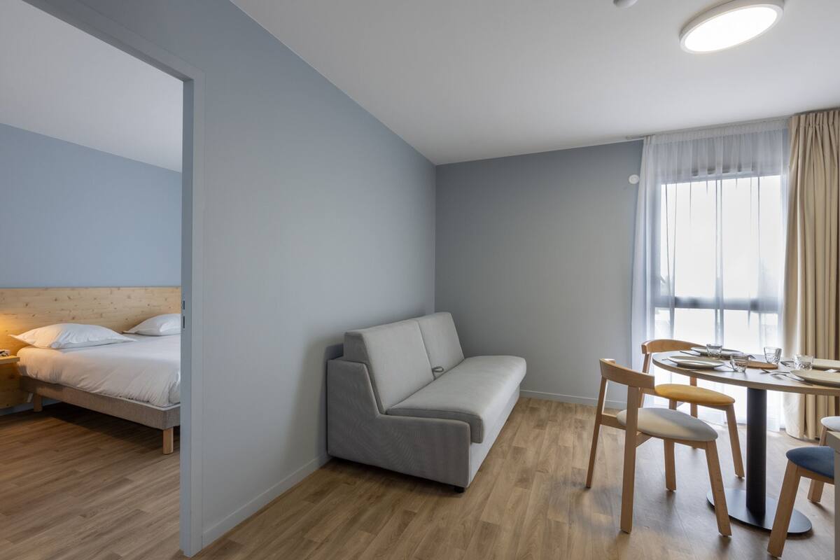 A modern hotel room with a bed, a sofa, a dining table with chairs, and a window with curtains.