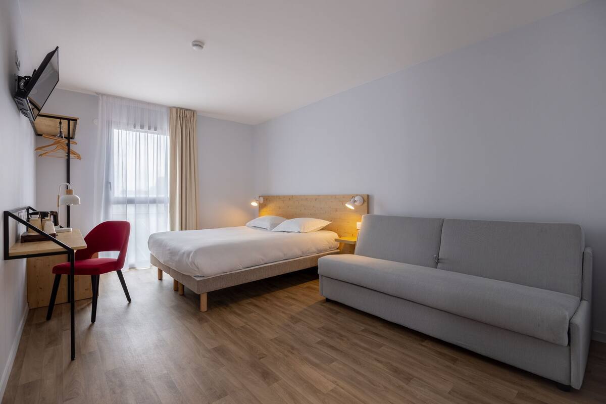 A modern hotel room with a bed, a grey sofa, a desk with a chair, and a TV mounted on the wall.