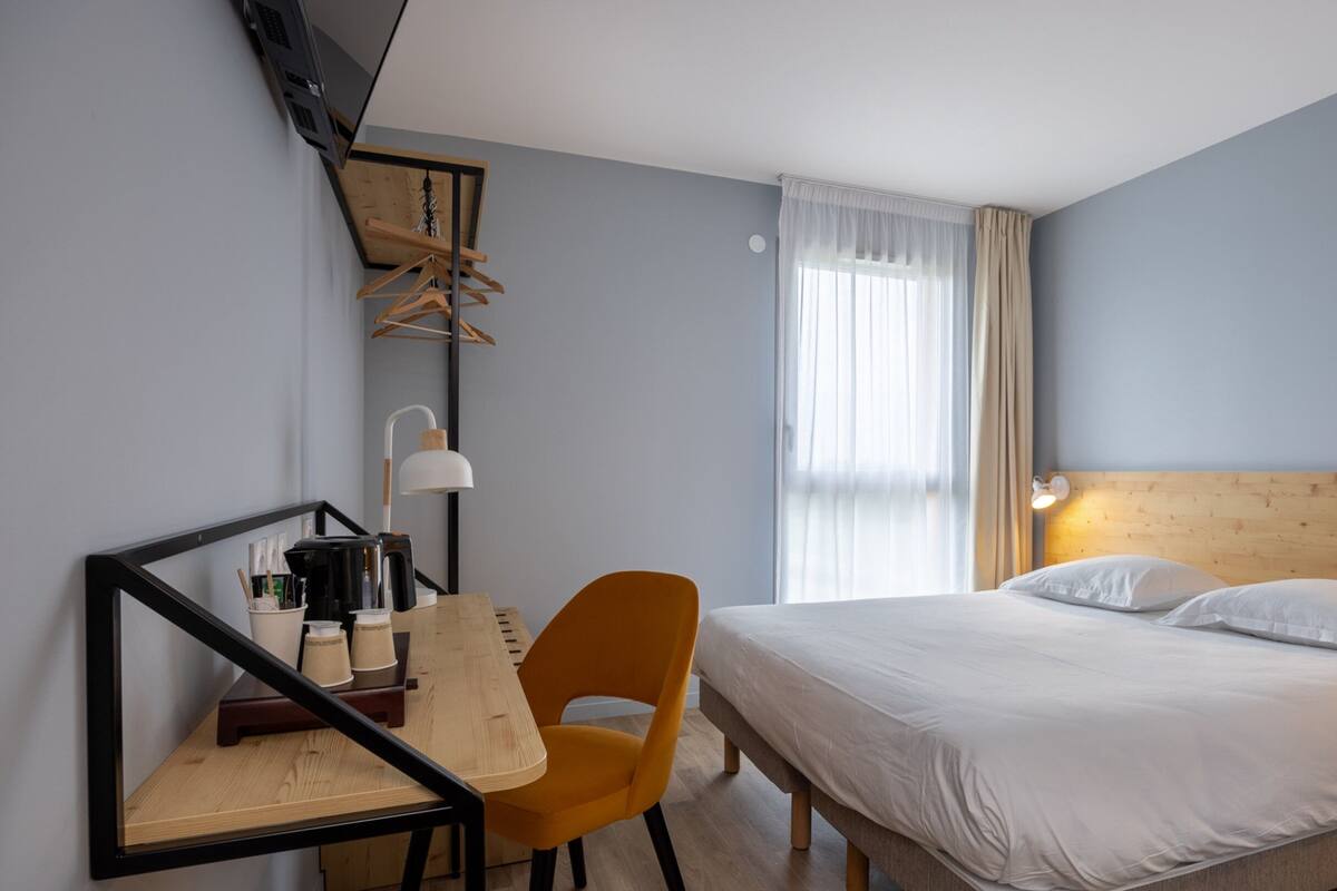 A hotel room with a bed, a desk with a chair, a mirror, and a window with curtains.