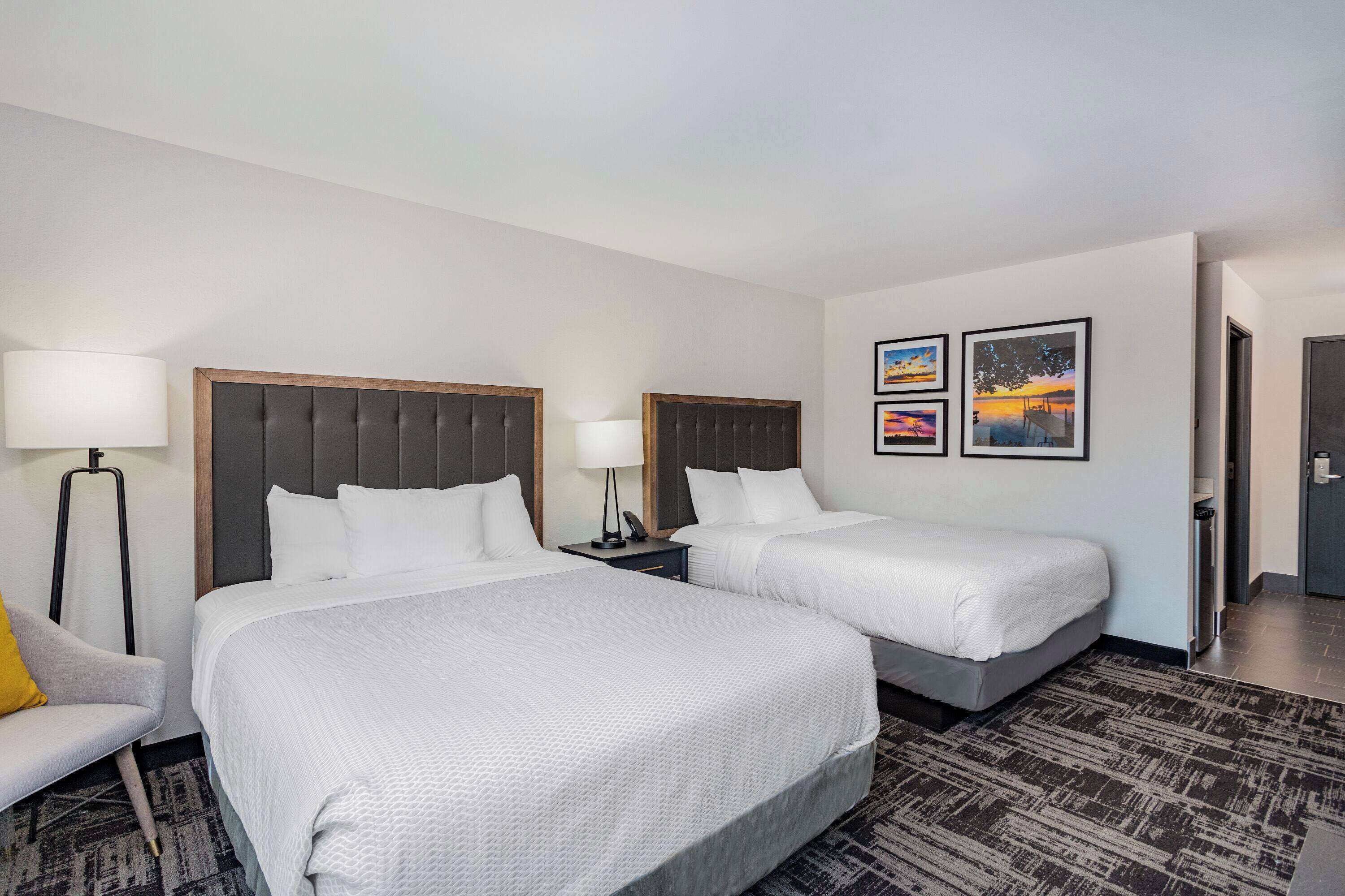 A hotel room with two beds, a desk, a chair, and a lamp. There are two framed pictures on the wall.