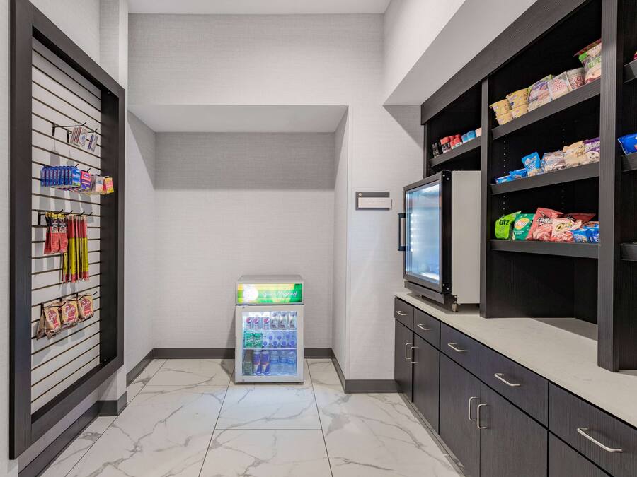 A compact hotel room with a minibar, a small refrigerator, and a shelf stocked with snacks.