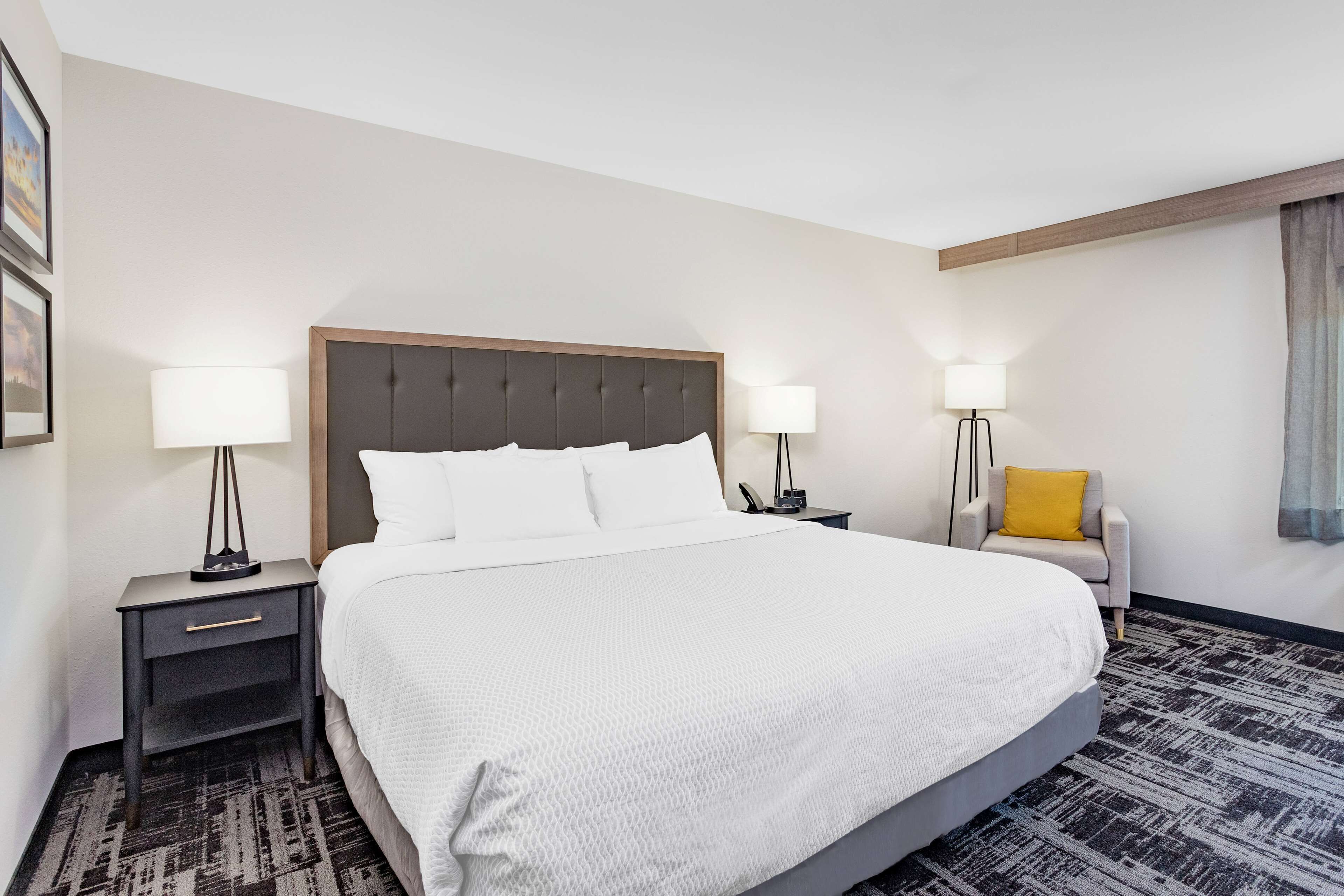 A hotel room with a large bed, two bedside tables with lamps, a chair with a yellow cushion, and a patterned carpet.