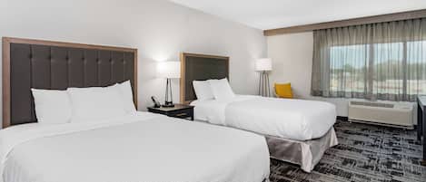 Habitación de hotel con dos camas, un escritorio, una silla, una lámpara y un ventanal con cortinas.