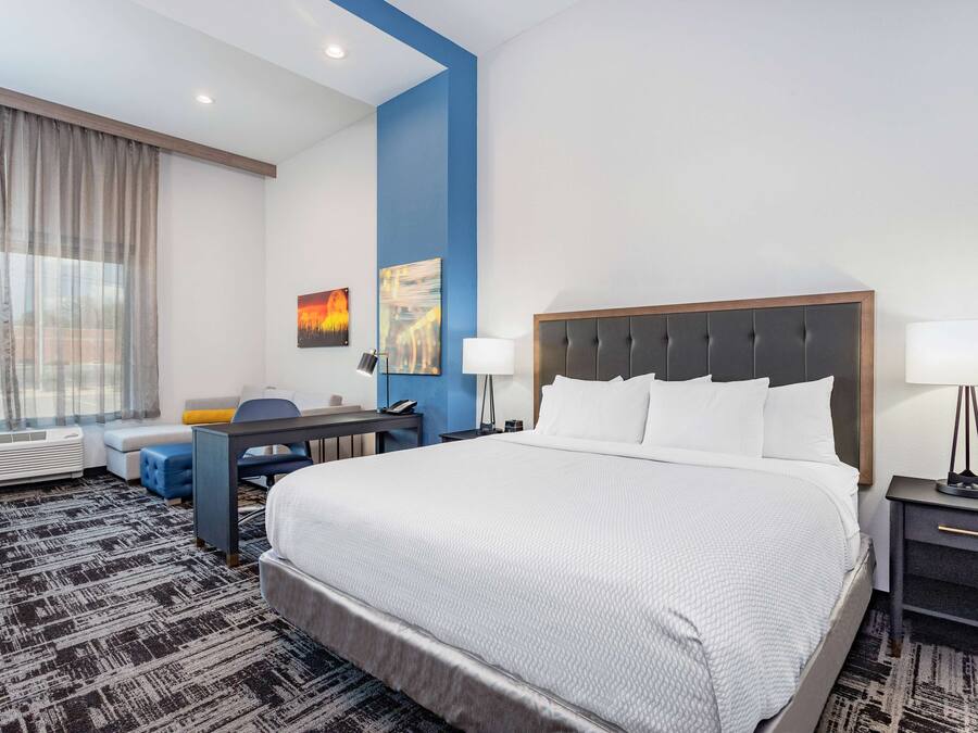 Habitación de hotel con una cama grande, un escritorio, una silla, una lámpara y un ventanal con cortinas.