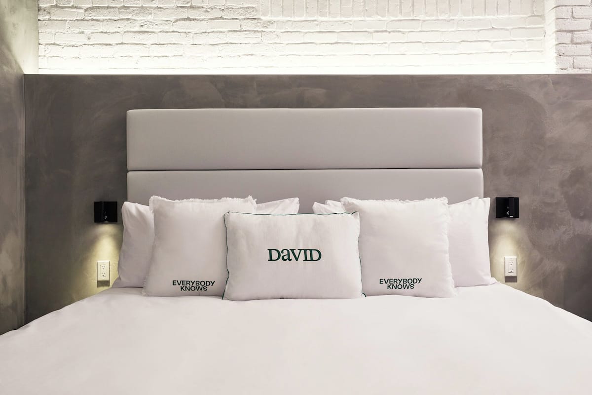 Una cama bien hecha con cabecera, tres almohadas y dos luces de noche fijadas a la pared.