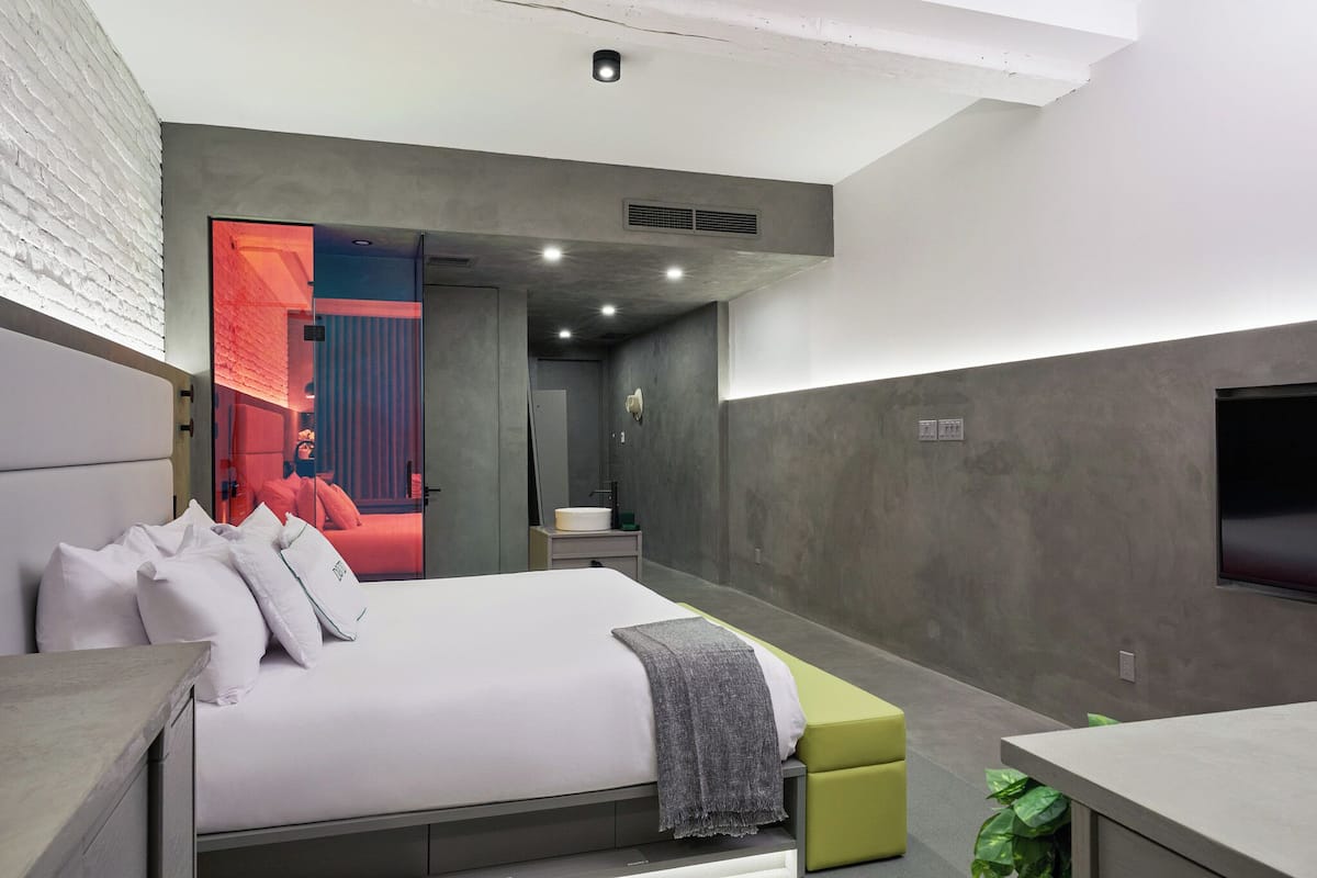 Un dormitorio moderno con una cama grande, una mesita de noche, un banco verde y un baño visible a través de una puerta abierta.