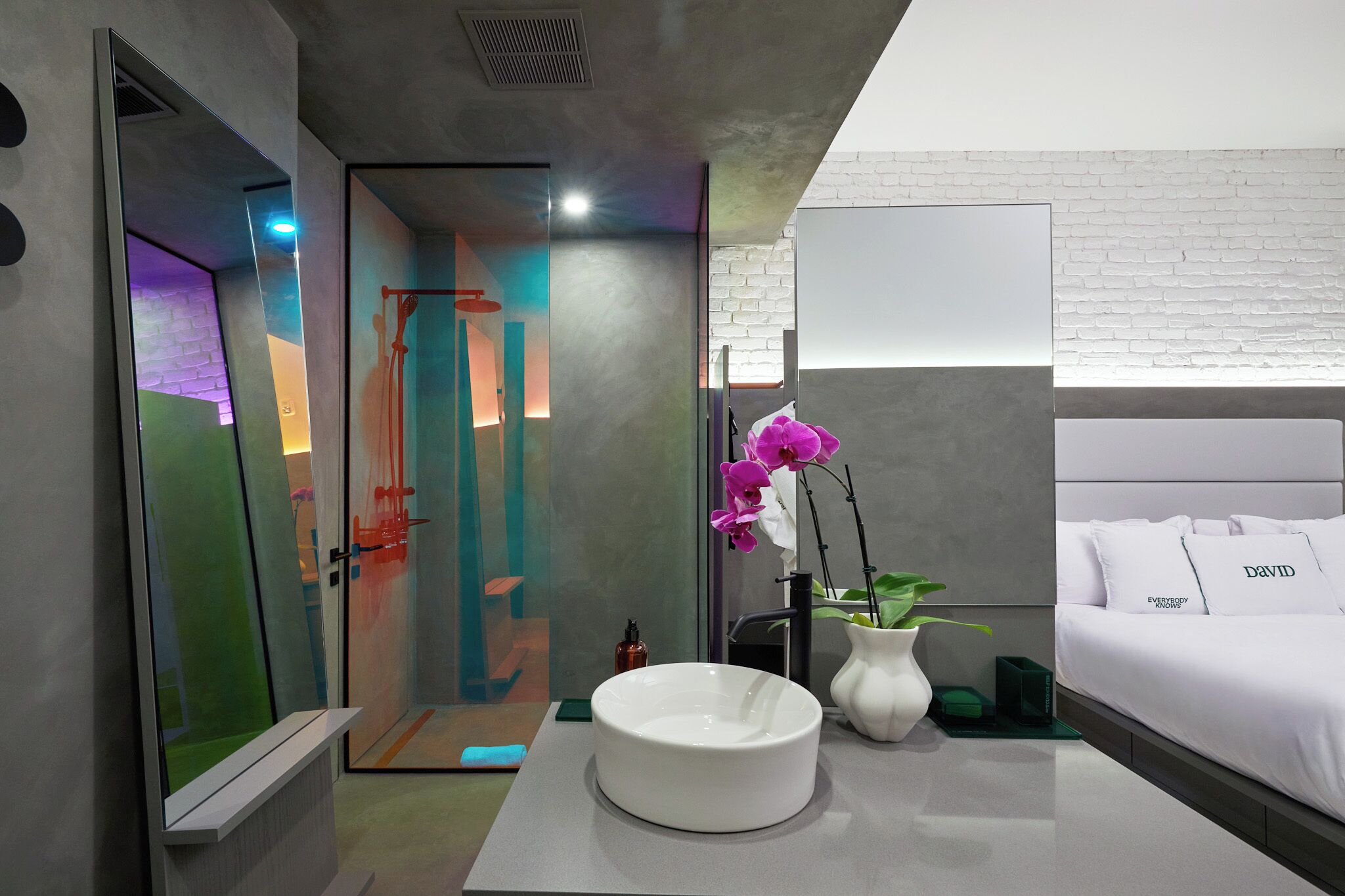 Una habitación de hotel moderna con ducha de vidrio, una cama blanca con un cojín que dice 'DAVID' y un jarrón con una planta.