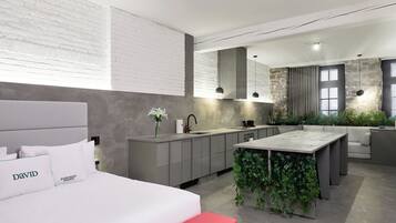 A modern hotel room with a bed, a small island with greenery, a kitchenette, and a seating area with a couch.