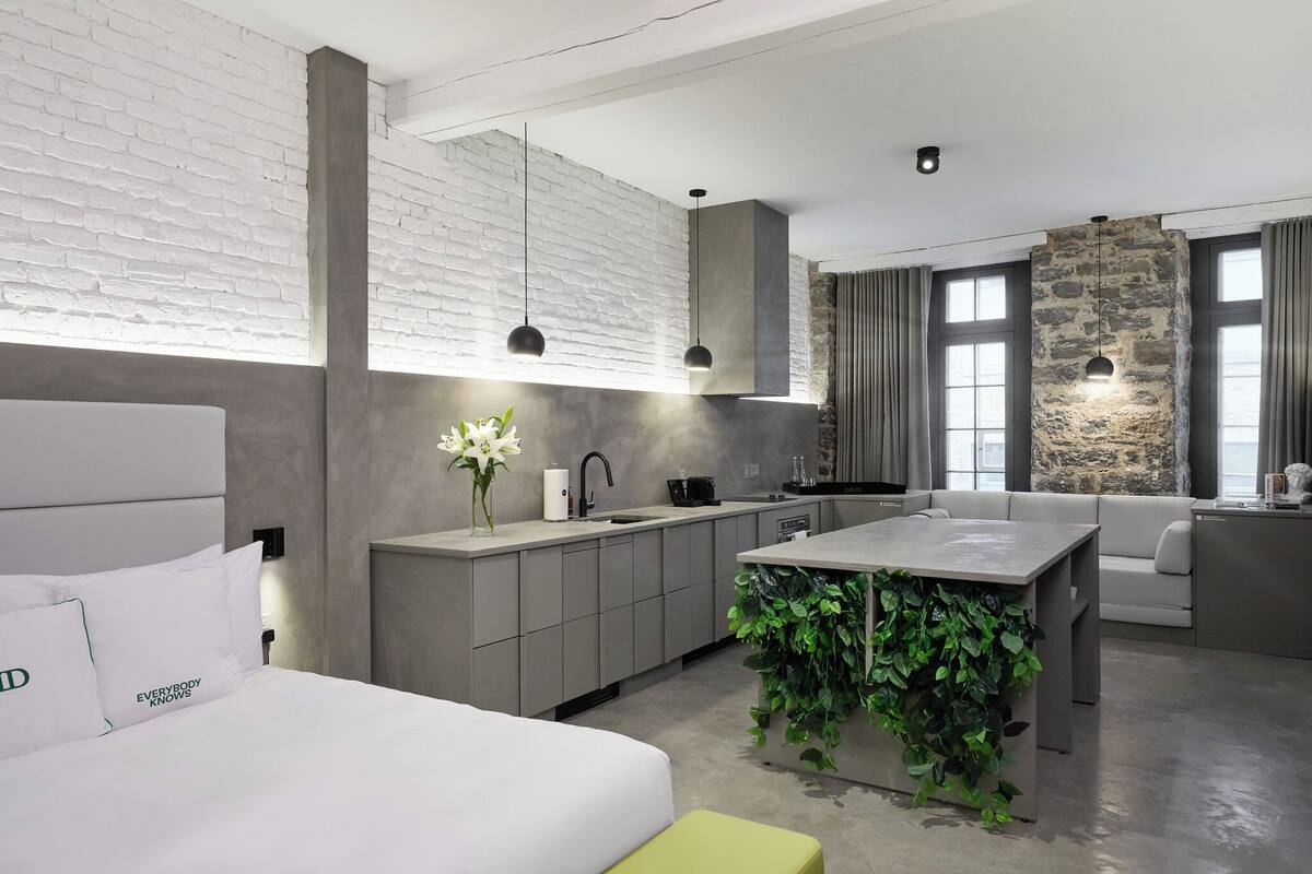 Una habitación de hotel moderna con una cama grande, un kitchenette, un comedor y una sala de estar con una pared de acento de piedra y ventanas amplias.
