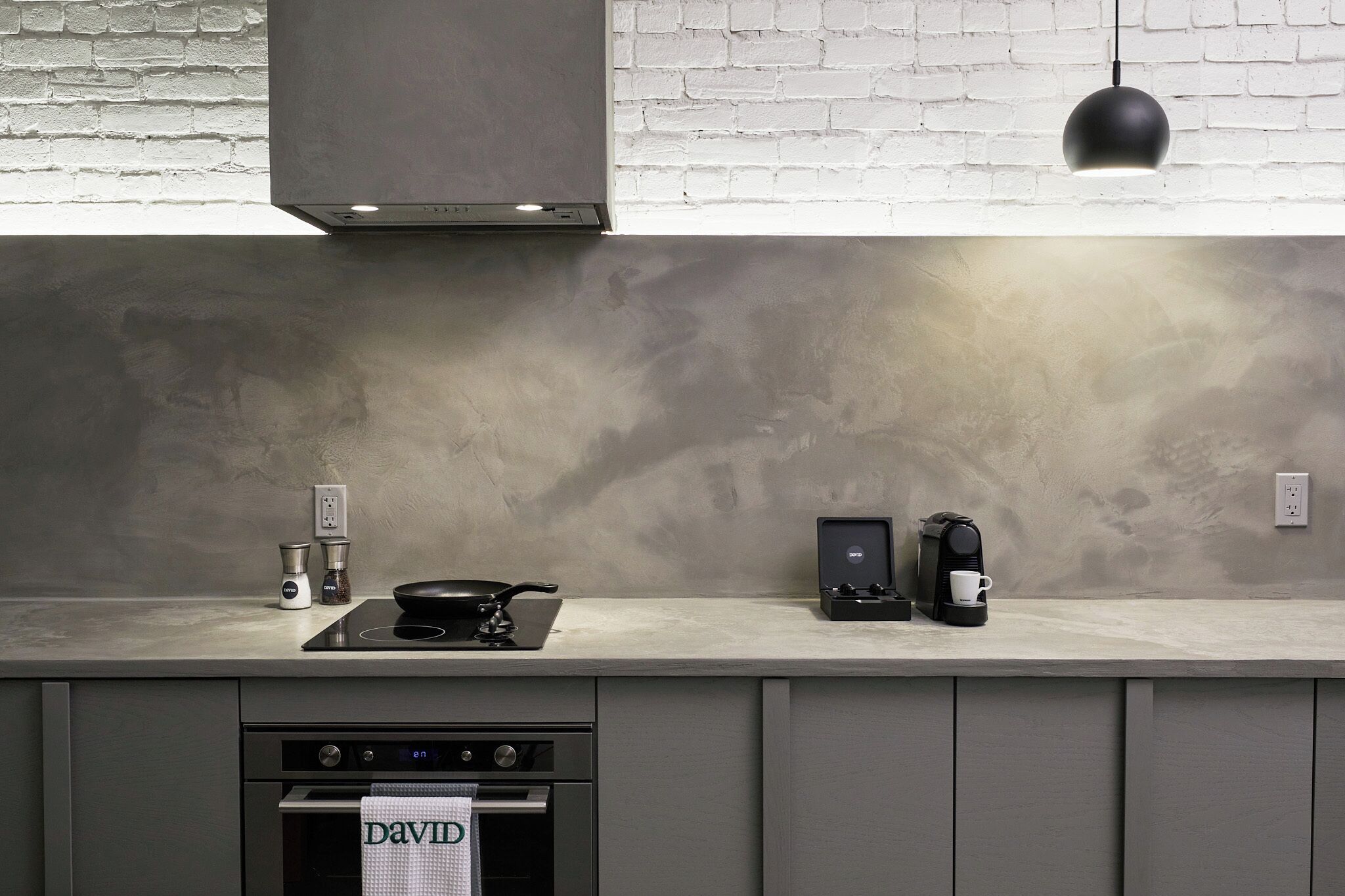 A modern kitchen with a brick wall, a countertop with a coffee maker, a kettle, and a frying pan.